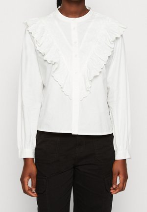 Blouse blanche à manches longues avec un col en dentelle volante et un devant à boutons, portée avec un pantalon noir.