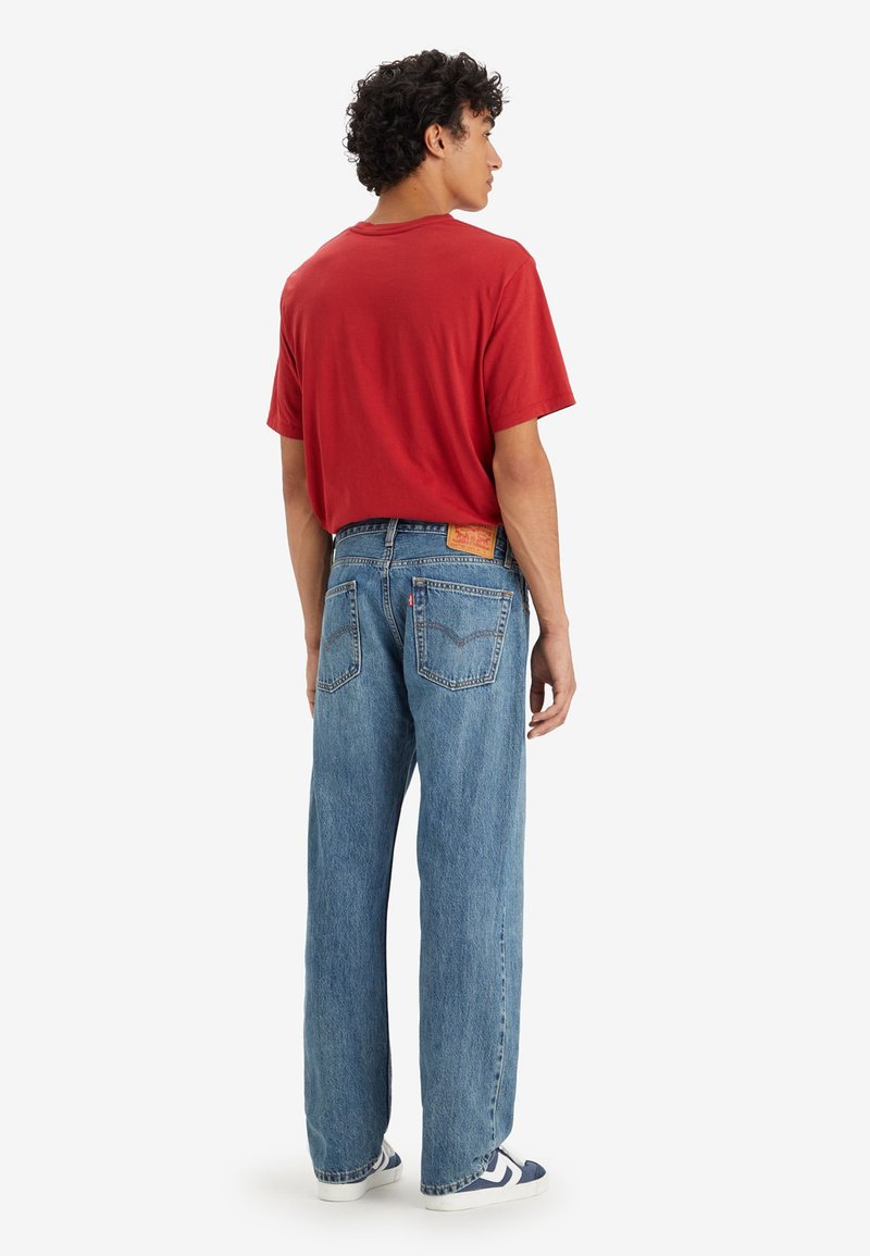 Camiseta de algodón roja, ajuste holgado. Jeans de mezclilla azul, corte ancho, cinco bolsillos, parche de marca visible en la cintura trasera. Zapatillas blancas y azules.