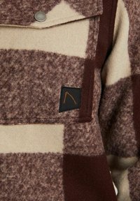 Fleece shirt met een bruin en crèmekleurig geruit patroon, drukknop op de borstzak en een zwart label met een oranje embleem.