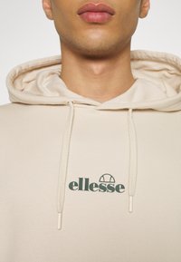 Beżowa bluza z kapturem z przodu z logo "ellesse" w ciemnozielonym kolorze. Posiada kaptur ze sznurkiem i fakturowaną tkaninę. Prosty, casualowy design.