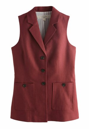 Kvinde står på terrasse ved havet iført ærmeløs bordeaux blazer og matchende shorts, store hoop-øreringe og med sat håret tilbage.