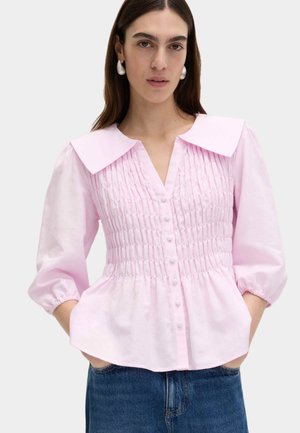 Donna che indossa una camicetta rosa chiaro con bottoni, colletto ampio, corpetto arricciato, maniche a sbuffo lunghe fino a metà braccio e jeans blu, con le mani nelle tasche.