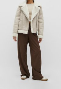 Světlá béžová bunda ze srsti s širokým límcem, zapínáním na zip a dvěma kapsami, kombinovaná s volnými hnědými kalhotami a teniskami.