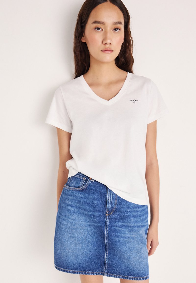Λευκό βαμβακερό T-shirt με V-λαιμόκοψη και κοντά μανίκια, με μικρό λογότυπο στο αριστερό στήθος. Συνδυάζεται με μπλε τζιν φούστα.