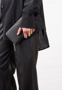 Portefeuille en cuir noir avec une texture lisse, fermeture éclair et matériel en argent, tenu dans une main, associé à des vêtements noirs.