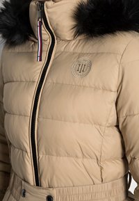 Béžová prošívaná bunda s kapucí lemovanou černým umělým kožíškem, předním zipem a páskem na zavazování. Má polstrované části a nášivku s logem na hrudi.