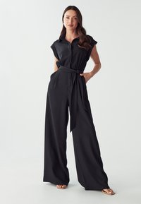Zwarte jumpsuit met korte cap mouwen, knoopsluiting aan de voorkant, ceintuur in de taille, wijde pijpen, gladde textuur en zijzakken.