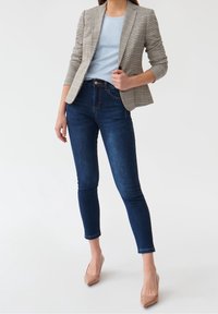 Bleiseri vaaleassa ruudullisessa kankaassa, sininen paita, tummat kireät farkut ja nudenväriset korkokengät. Muotoutuva istuvuus, kontrastiväriset ompeleet, lyhyet hihat.