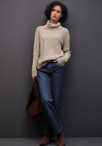 Suéter de punto beige con un cuello alto, patrón texturizado; pantalones vaqueros azul oscuro; botas de ante marrón hasta el tobillo; sosteniendo un bolso marrón.