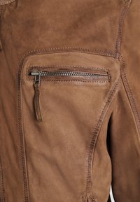 Sezione di giacca in suede marrone che mostra una tasca orizzontale con zip e un tirante in pelle, con cuciture dettagliate.