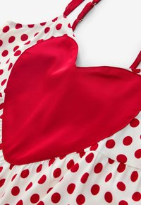 Biele šaty s červenými bodkami, s veľkou červenou saténovou aplikáciou v tvare srdca na živôtiku a nastaviteľnými ramienkami na prispôsobenie.