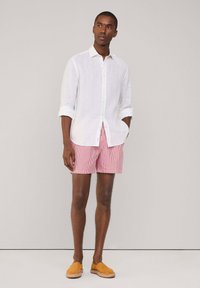 Witte linnen blouse, opgerolde mouwen; rood-witte gestreepte shorts; gele schoeisel; neutrale achtergrond.