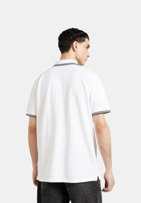 Polo bianco con colletto strutturato e strisce blu navy lungo il colletto e le maniche. Realizzato in un tessuto liscio, presenta un orlo dritto.