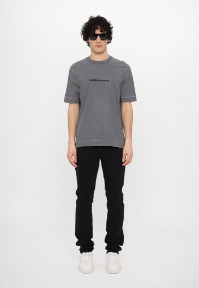 T-shirt cinza riscada com logótipo preto, combinada com calças pretas e ténis brancos. O modelo usa óculos de sol pretos. Conjunto simples e casual.