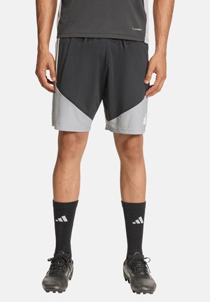 Mann trägt schwarze und graue Sportshorts, schwarze Socken mit weißem Logo und schwarze Fußballschuhe, steht vor weißem Hintergrund.