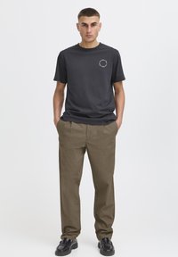 T-shirt noir en coton avec col rond et logo à l'avant, associé à un pantalon kaki à coupe décontractée et des chaussures noires, posé devant un fond uni.