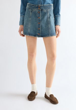 Personne portant une jupe en denim bleue boutonnée, un haut à manches longues en denim bleu, des chaussettes blanches et des mocassins marron, debout sur un fond blanc.
