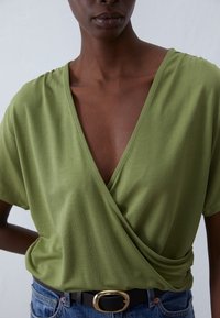 Olive green v-neck wrap top in soft fabric with short sleeves, paired with denim jeans and a black belt featuring a gold buckle.