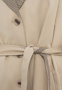 Trench-coat beige avec un revers en pied-de-poule, deux boutons et une ceinture nouée assortie. Texture lisse et coupe cintrée.