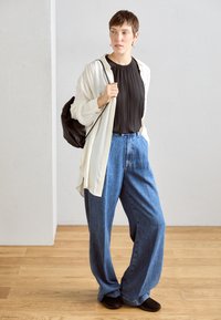Haut plissé noir, chemise à boutons crème surdimensionnée, jeans en denim bleus à jambes larges et chaussures noires ; accessoires subtils exposés.