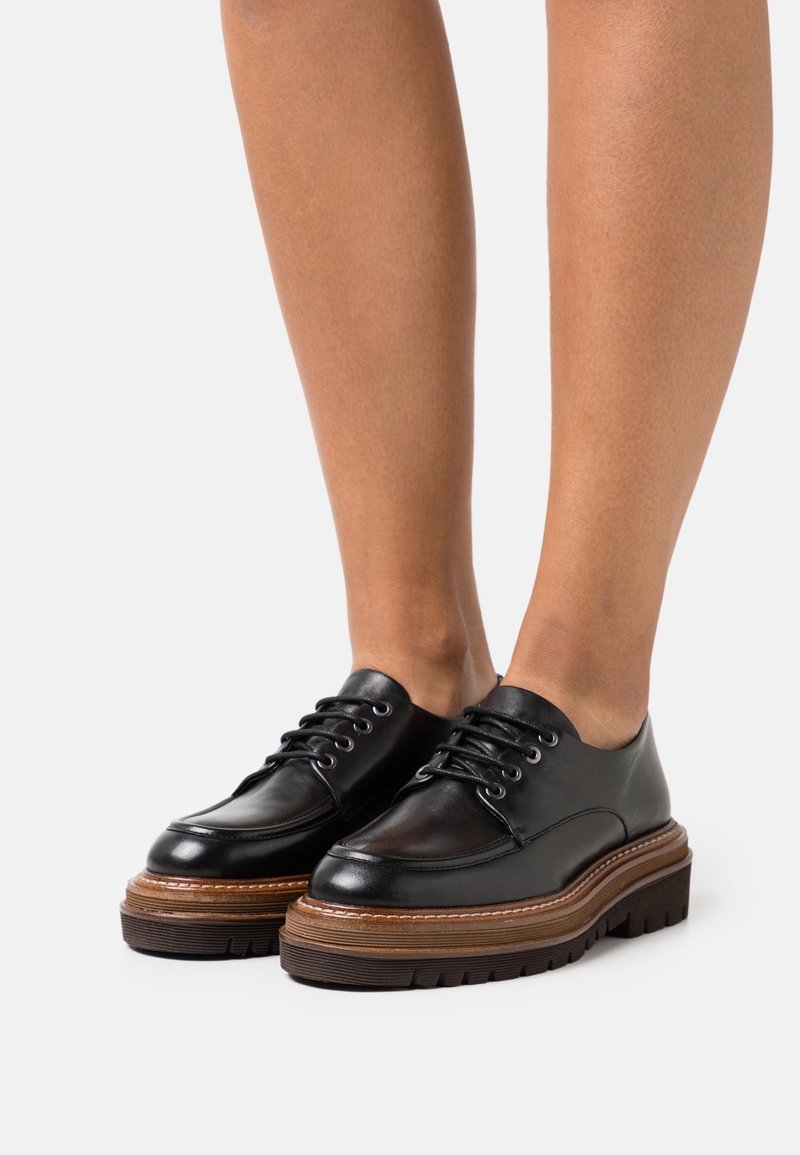 Chaussures à plateforme en cuir noir à lacets portées pieds nus, avec des semelles épaisses texturées marron et noires, présentées sur un fond uni.