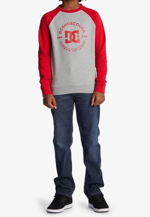 Grauer Sweatshirt mit roten Ärmeln, Logo in der Mitte, gerippte Bündchen und Saum, kombiniert mit blauen Jeans und schwarzen Sneakern. Lässiges, sportliches Design.