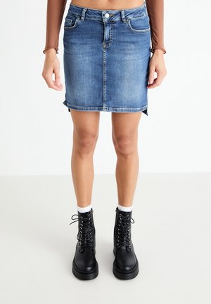 Persoon die een blauwe denim minirok draagt, zwarte veterlaarsjes tot aan de enkel, witte sokken en een cropped bruin langärmelig topje tegen een effen achtergrond.