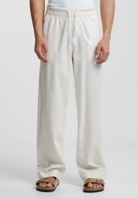 Linnebyxor i ljus krämfärg med elastisk midja och avslappnad passform, med en slät textur och vida ben. Sandaler på fötterna.