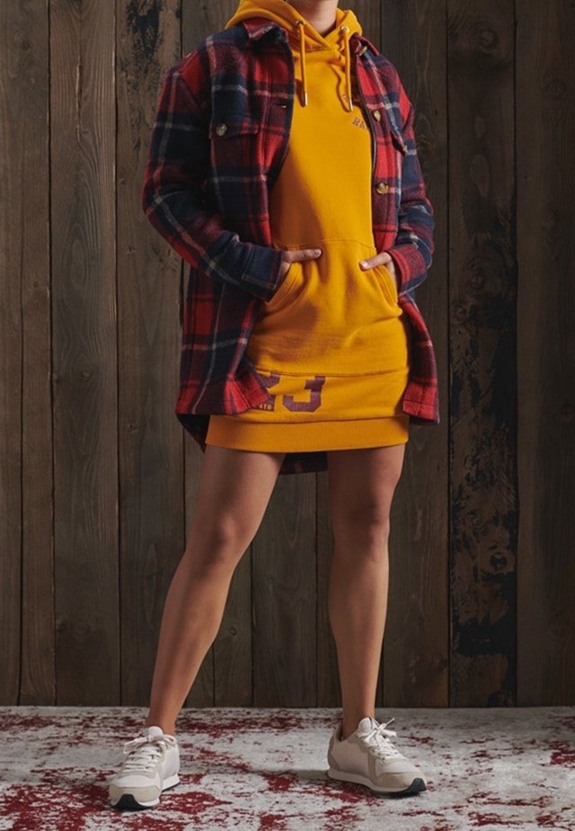 Personne debout les mains dans les poches d'une robe à capuche jaune, portant une veste à carreaux rouge et bleu marine et des baskets blanches sur un sol en bois.