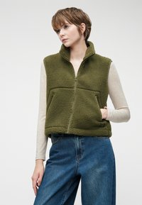 Zelena flis vetrovka z visokim ovratnikom, sprednjim zadrgo in dvema žepoma. Nosi se čez svetlo majico z dolgimi rokavi, v kombinaciji z ohlapnimi modrimi kavbojkami.