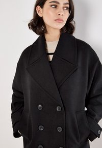 Cappotto nero doppiopetto in un tessuto morbido, caratterizzato da grandi bottoni e tasche laterali, con un ampio colletto e una vestibilità rilassata.