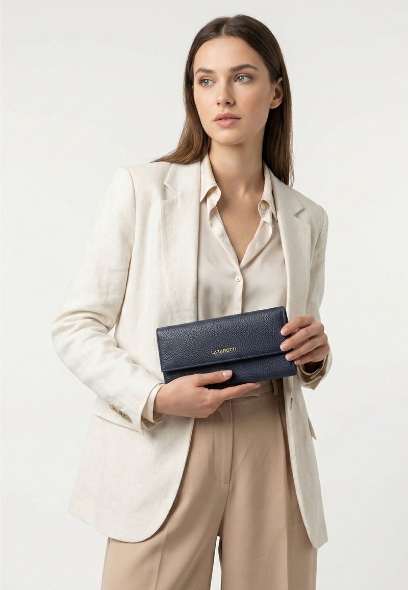 Mujer con blazer y pantalones beige sostiene con ambas manos una cartera Lazarotti azul marino sobre un fondo liso y claro.