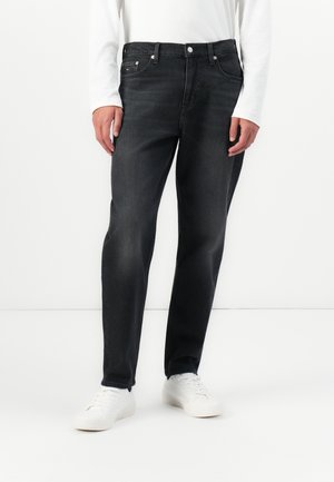 Man wearing black slim-fit jeans and white sneakers standing against a white background with hands relaxed by sides.