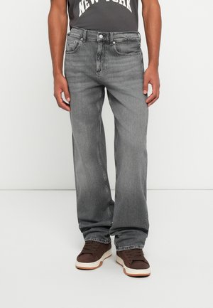 Mand iført gråslidte jeans med lige ben og brune sneakers står mod en hvid baggrund.