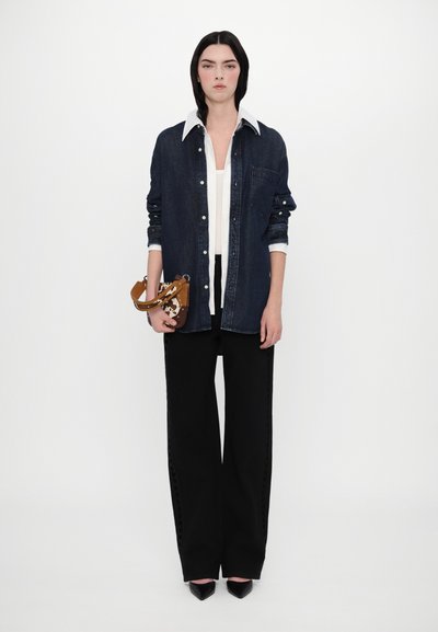 Chemise en denim avec un col blanc, associée à un top blanc et un pantalon large noir. Tient un sac à main à motifs avec une bandoulière marron.