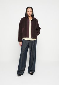 Weinrotes Fleecejacke mit hohem Kragen, Reißverschluss vorne und Seitentaschen, kombiniert mit lockeren, dunklen Denimhosen und einem hellen Oberteil.