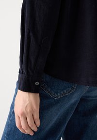 Gros plan d'une main reposant sur le côté, portant une chemise à manches longues foncée avec un poignet boutonné et un jean en denim bleu avec une poche arrière visible.