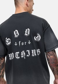 T-shirt noir surdimensionné avec un lettrage gothique blanc audacieux au dos, affichant un effet dégradé. Manches courtes et coupe décontractée.