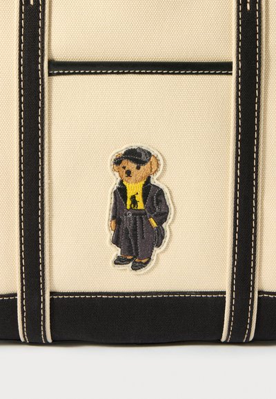 Saco de tela com acentos em preto. Apresenta um urso bordado a usar um suéter amarelo e um casaco preto, detalhado com costura em contraste.