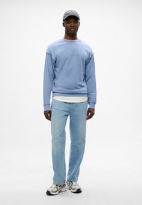 Sudadera azul claro con puños acanalados, que presenta un pequeño logo centrado. Combinada con jeans azul claro y zapatillas grises con acentos reflectantes.