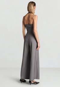 Donna che indossa una tuta grigia senza spalline a gambe larghe con scarpe nere basse e con punta, in piedi di schiena su uno sfondo semplice.