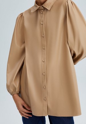 Femme portant un chemisier beige à manches longues boutonné, manches bouffantes et poignets élastiques, assorti à un pantalon bleu foncé.