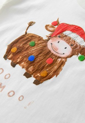 Valge puuvillane T-särk, millel on pruun, karvane lehma graafika, kes kannab punast jõuluvana mütsi, millel on erksad pom-pomid ja tekst all.