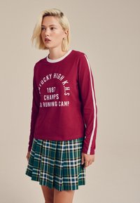 Punainen pitkähihaipaita, jossa valkoinen painatus ja raidat, yhdistetty vihreään ruudulliseen laskostettuun hameeseen, jossa on sinisiä, keltaisia ja valkoisia yksityiskohtia.