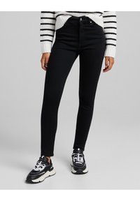 Svarta högmidjade skinny jeans i ett stretchigt denim-material, kombinerat med svartvita sneakers. En randig vit och svart tröja synlig.