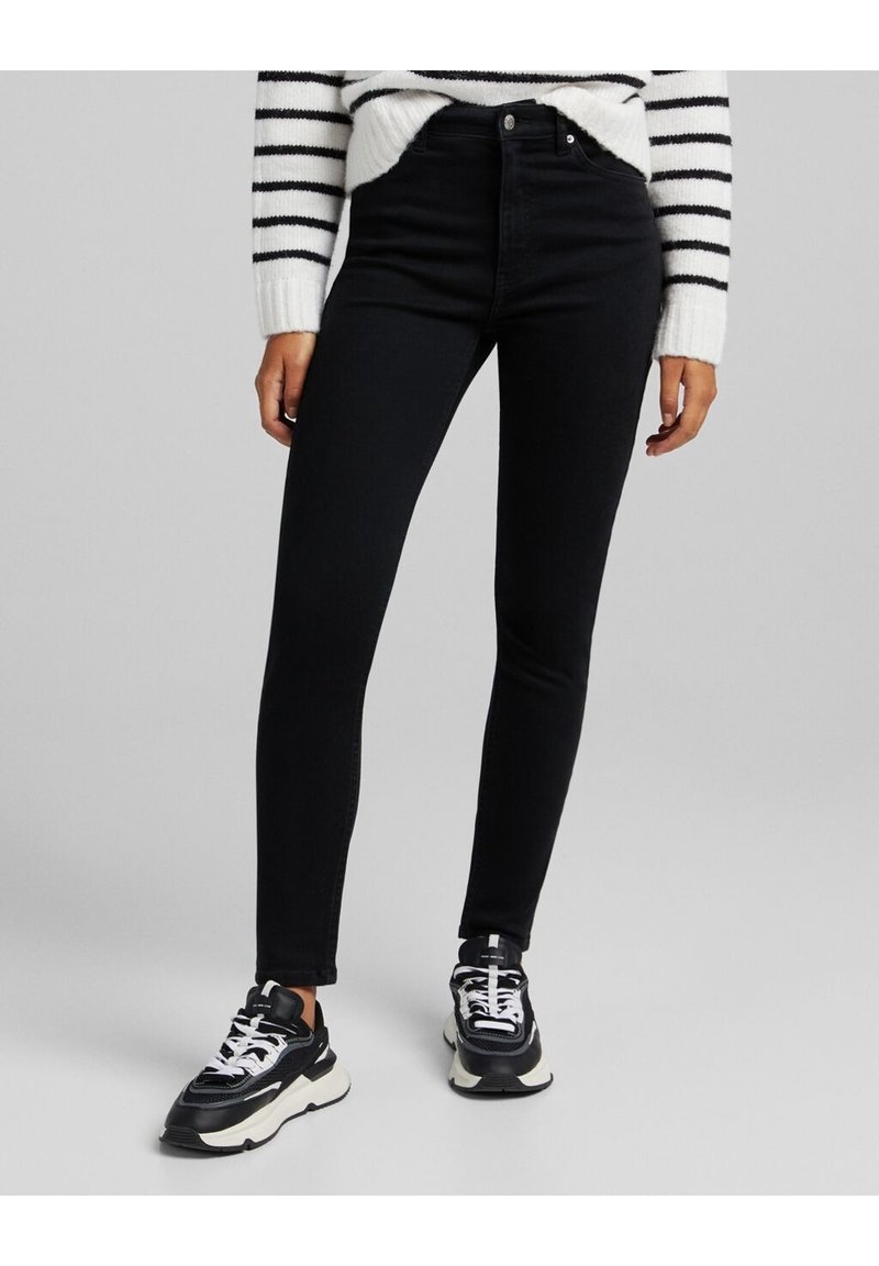 Svarta högmidjade skinny jeans i ett stretchigt denim-material, kombinerat med svartvita sneakers. En randig vit och svart tröja synlig.