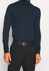 Pull à col roulé bleu marine en tissu doux, manches longues, coupe ajustée, porté avec un pantalon foncé et une ceinture en cuir marron à boucle argentée.