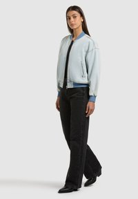 Femme debout portant une veste bomber bleu clair, un haut noir, un jean noir à jambes larges et des bottes noires à bout pointu, sur fond blanc.