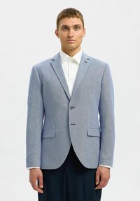 Blazer bleu clair en tissu texturé ; fermeture à un bouton, revers crantés et deux poches avant. Porté sur une chemise blanche.