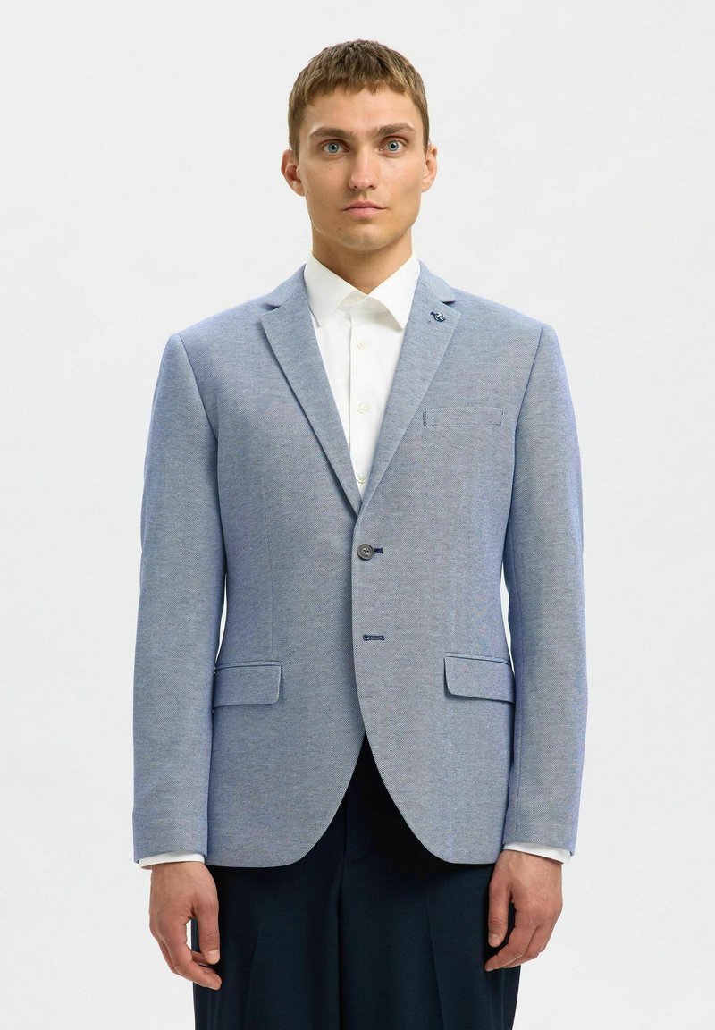 Blazer bleu clair en tissu texturé ; fermeture à un bouton, revers crantés et deux poches avant. Porté sur une chemise blanche.
